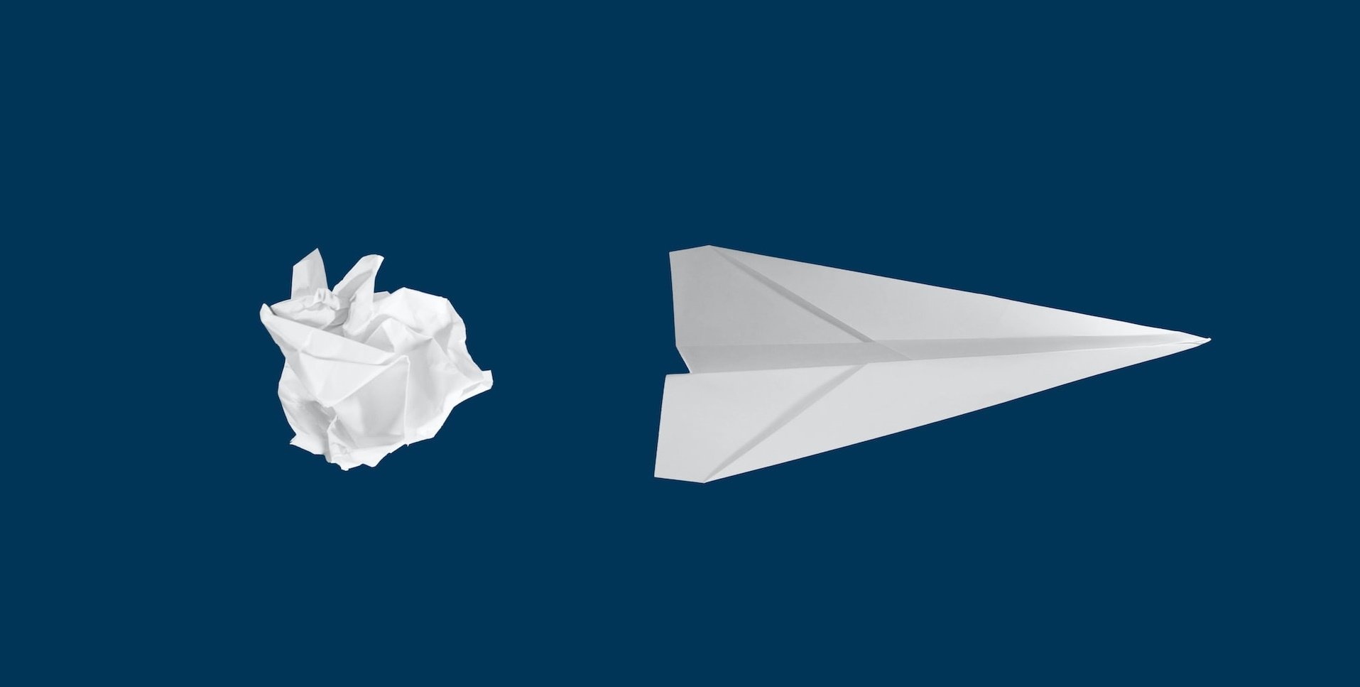 A scrunched up white paper to the left of a pristine white paper plane pointing right, on a blue background