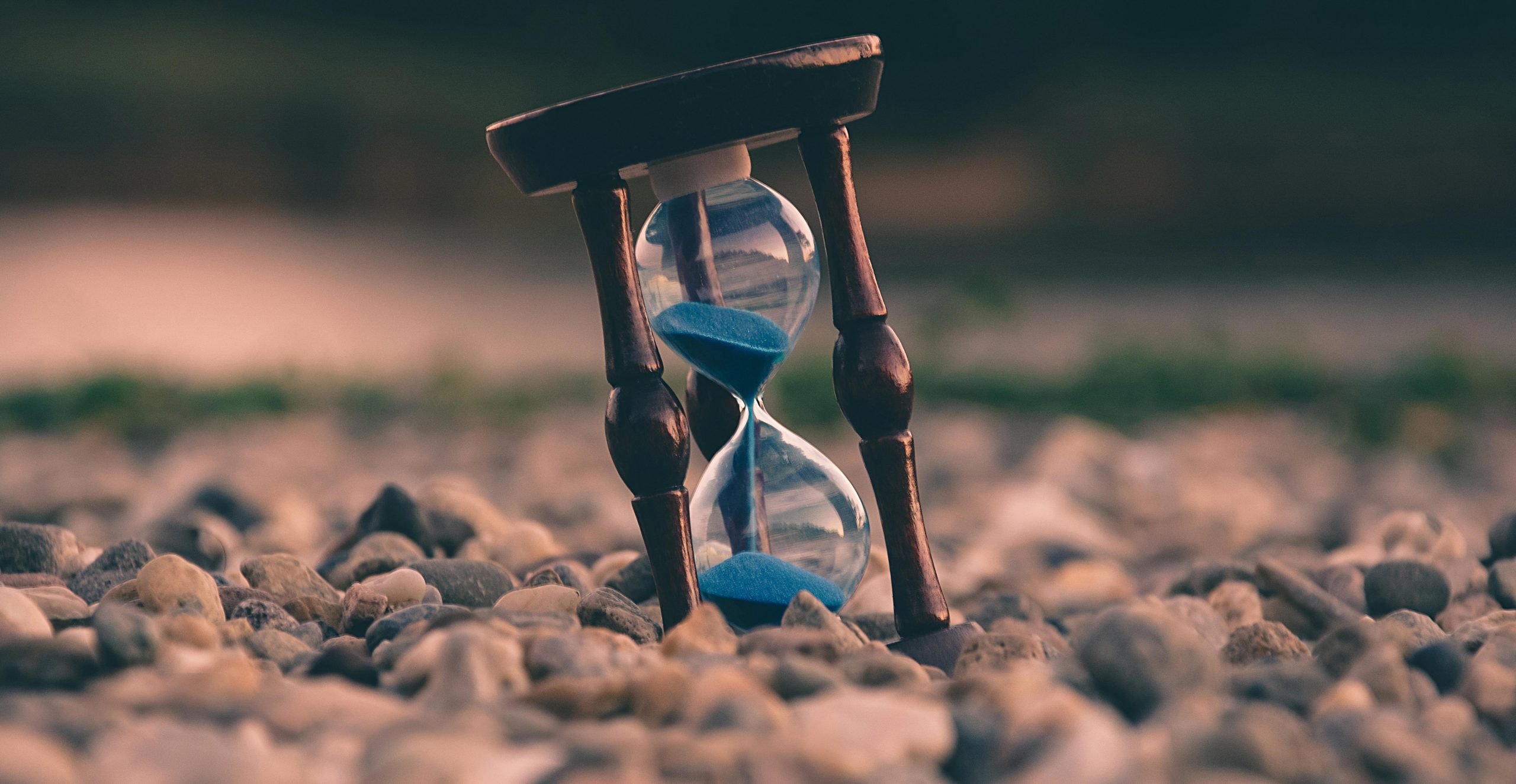 An hourglass with blue sand in it, perched on a pebble beach